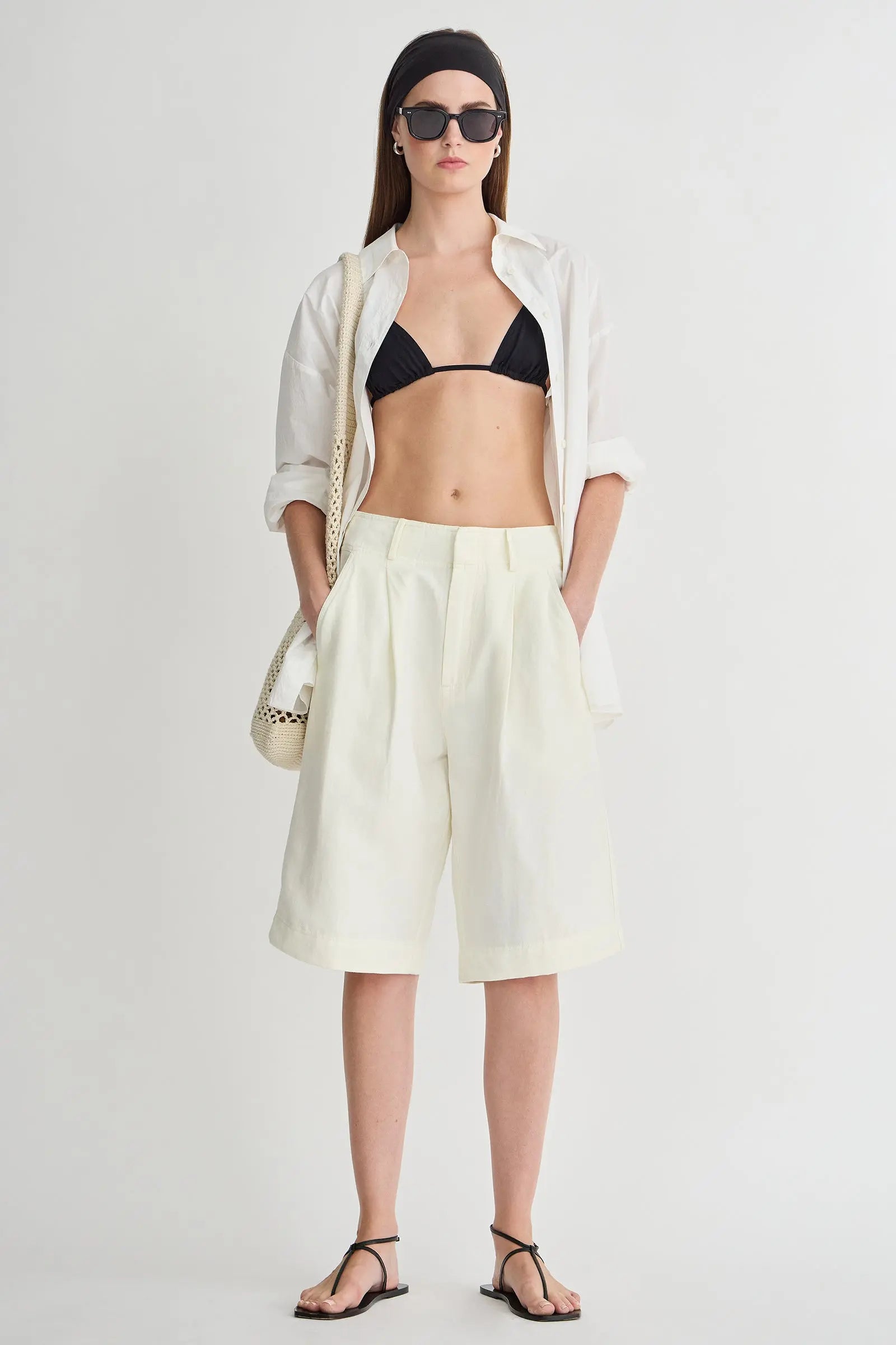 Woman wearing a white shirt, black bikini top, light yellow shorts, and black sandals on a white background