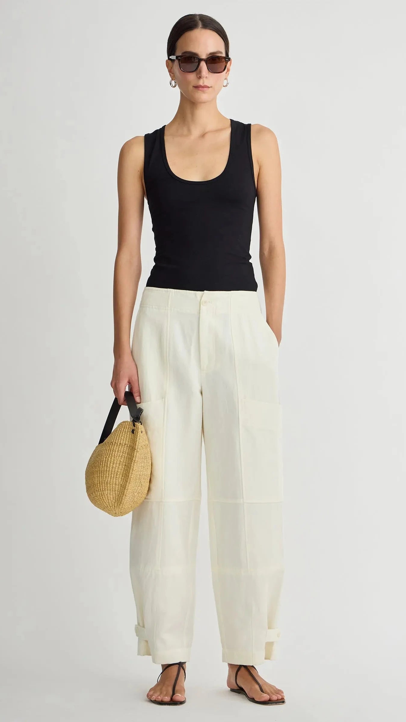 Woman wearing a black tank top and white pants, holding a straw bag on a light gray background