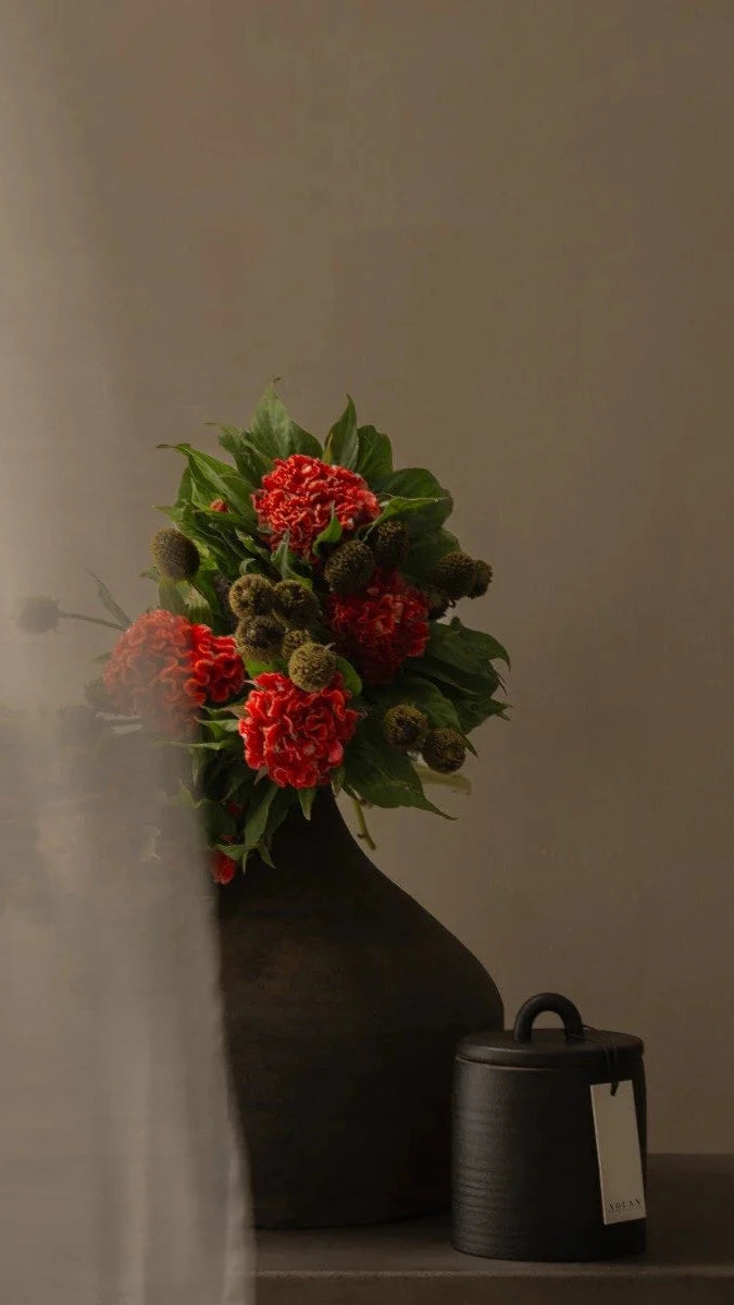 Black vase with red flowers and green leaves against a plain background
